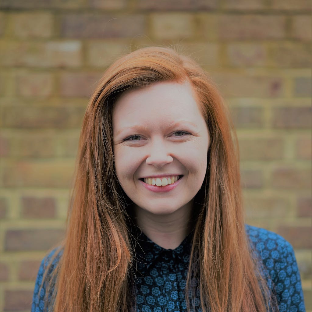 A headshot of Becky, Volunteering Development Manager. Becky is in front of a bricked wall and is wearing a blue top.