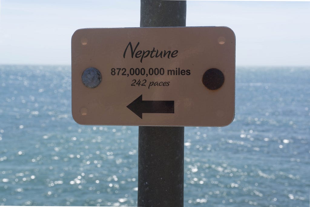 A small signpost with the sea in the background, reading ‘Neptune: 872,000,000 miles, 242 paces’.