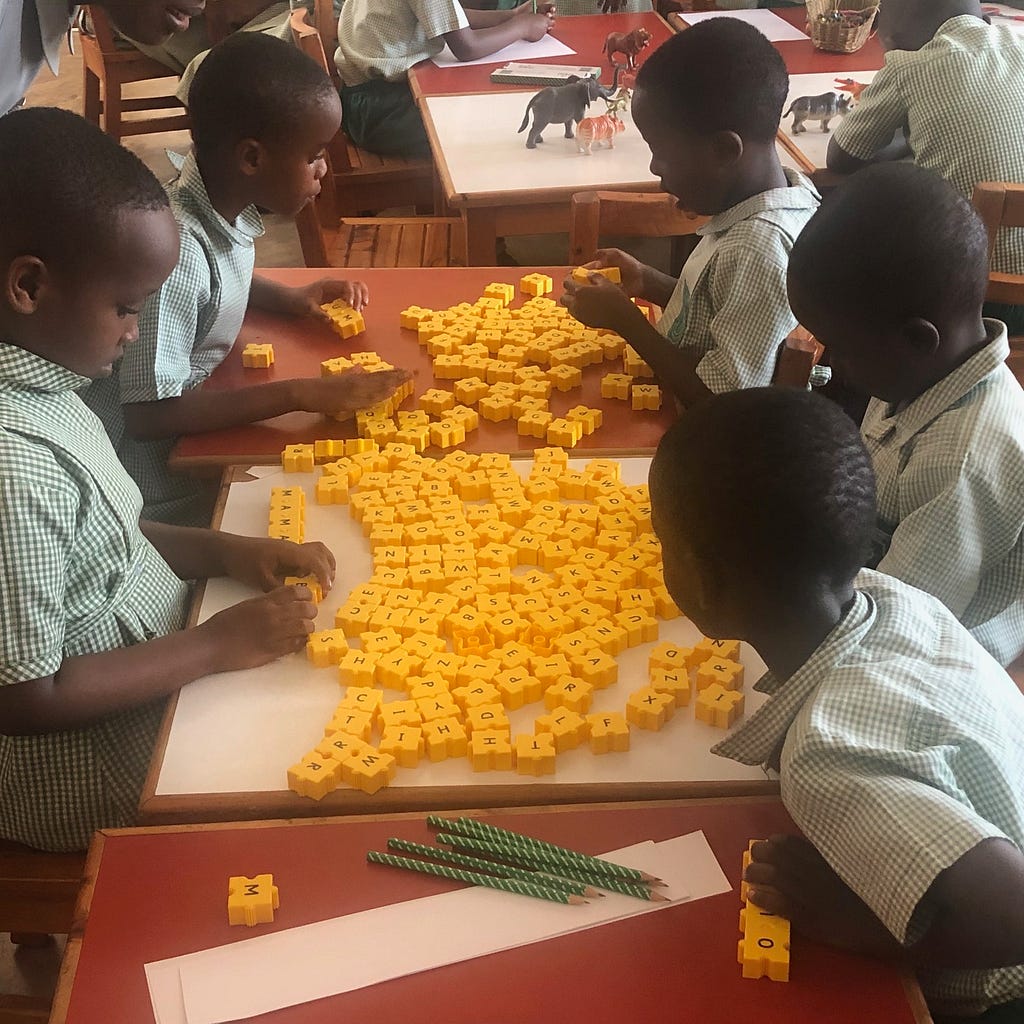 Five young children dressed in matching school uniforms handling bright yellow blocks — each printed with one letter of the alphabet — spread out over two school desks.