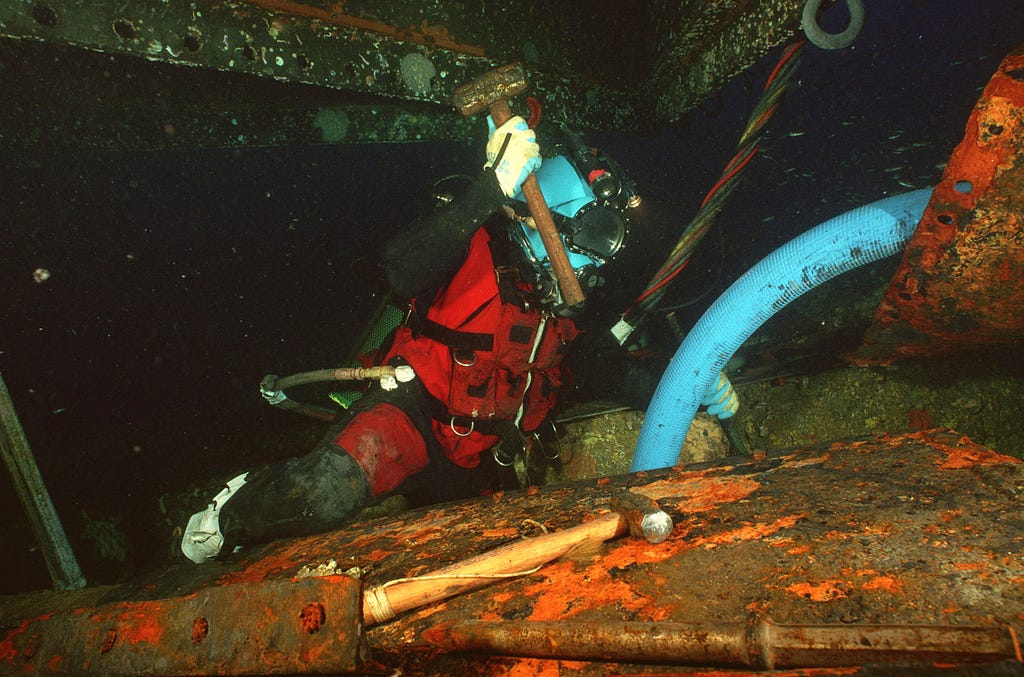 A diver holds a hammer poised above his head to chisel equipment.