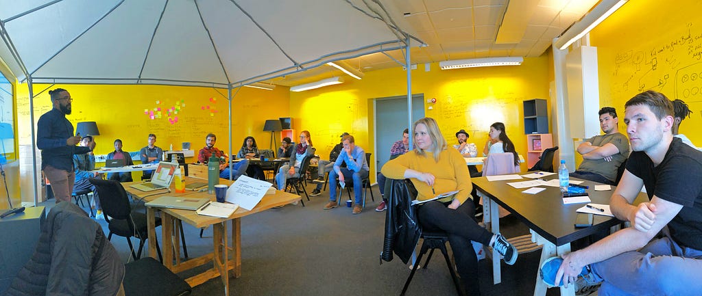 Eduardo presenting under a tent in the yellow room, students seated in a circle or chairs around him.