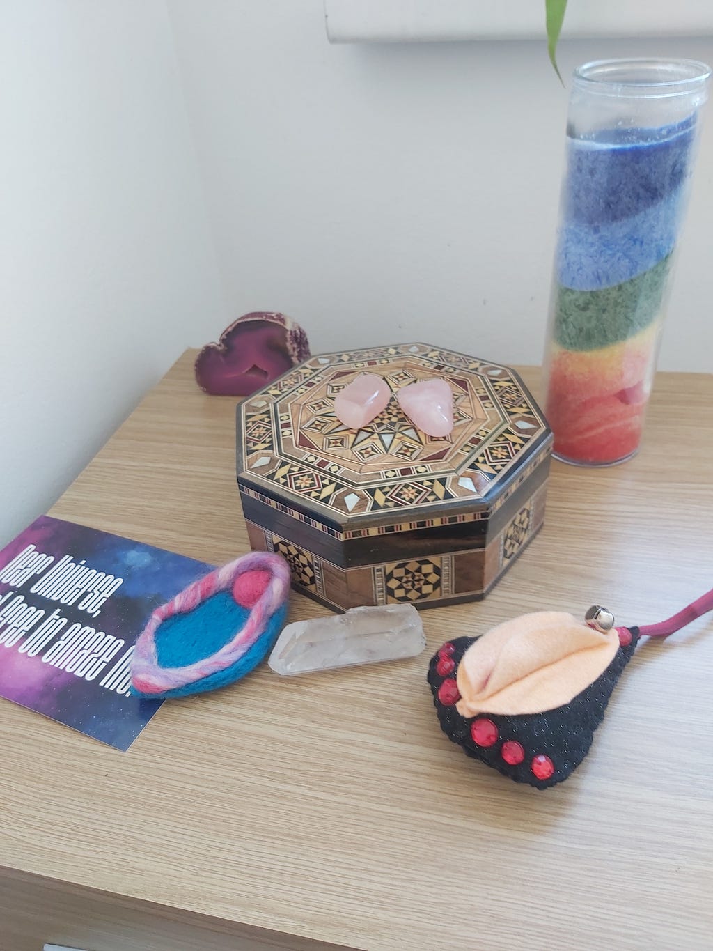 Picture of Lucy’s altar with a postcard saying, “Dear Universe, feel free to amaze me”, a blue and pink felted vulva, a clear quartz crystal, a black and peach vulva Christmas bauble, a gorgeous geometric wood box from Damascus, 2 rose quartz crystals on top, a pink crystal that Lucy can’t remember is but it looks sexy so we’re keeping it, and a rainbow candle