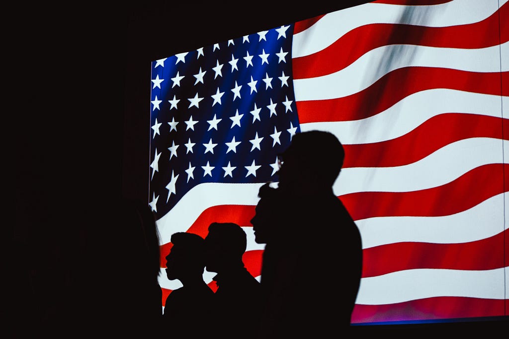 Dark silhouettes of people standing in front of the US flag.