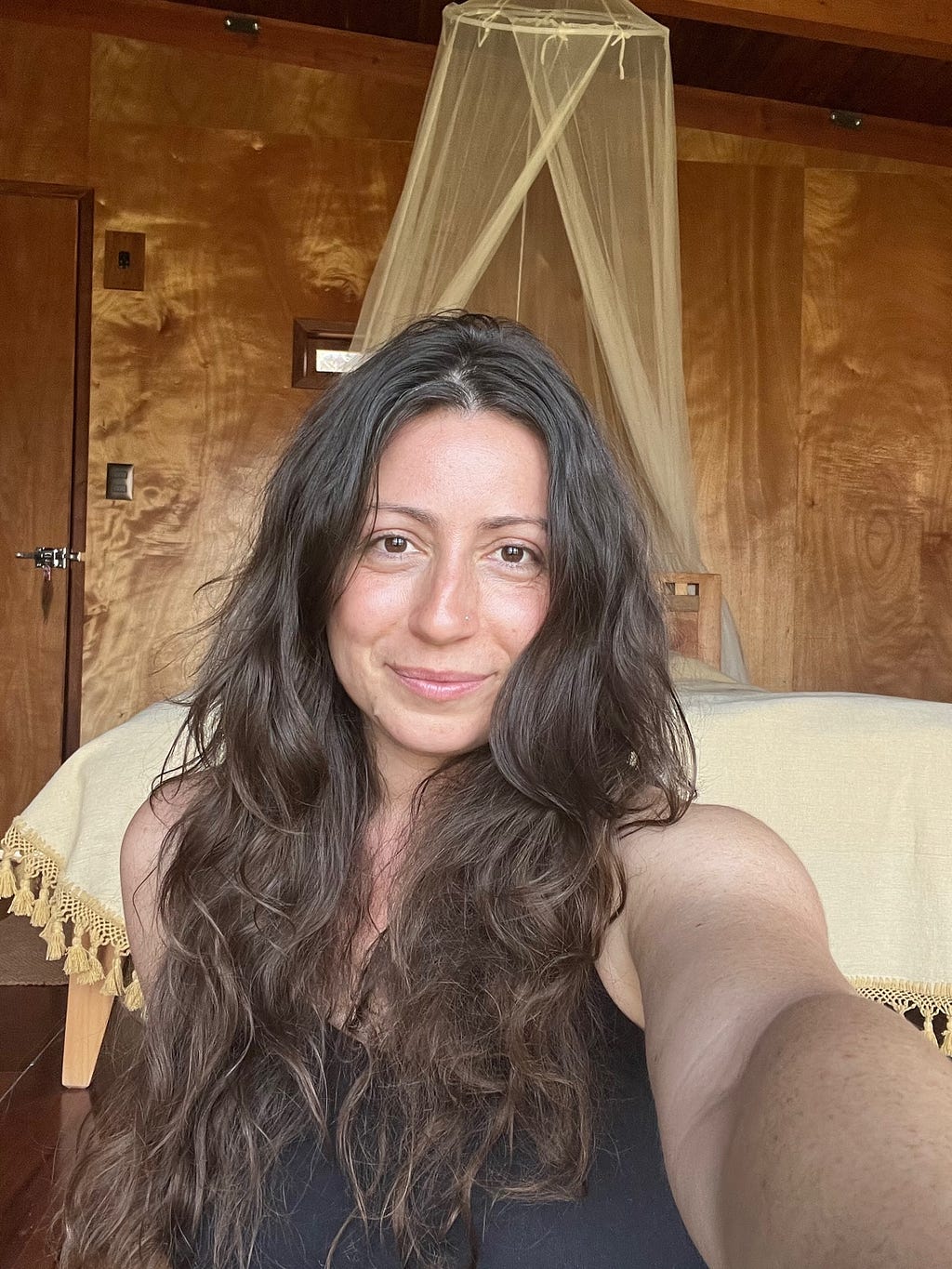 White woman with long brown hair, sun-browned skin smiling in a cabin. Shiny wood panels, a white mosquito net and a soft yellow bedspread are seen in the background.