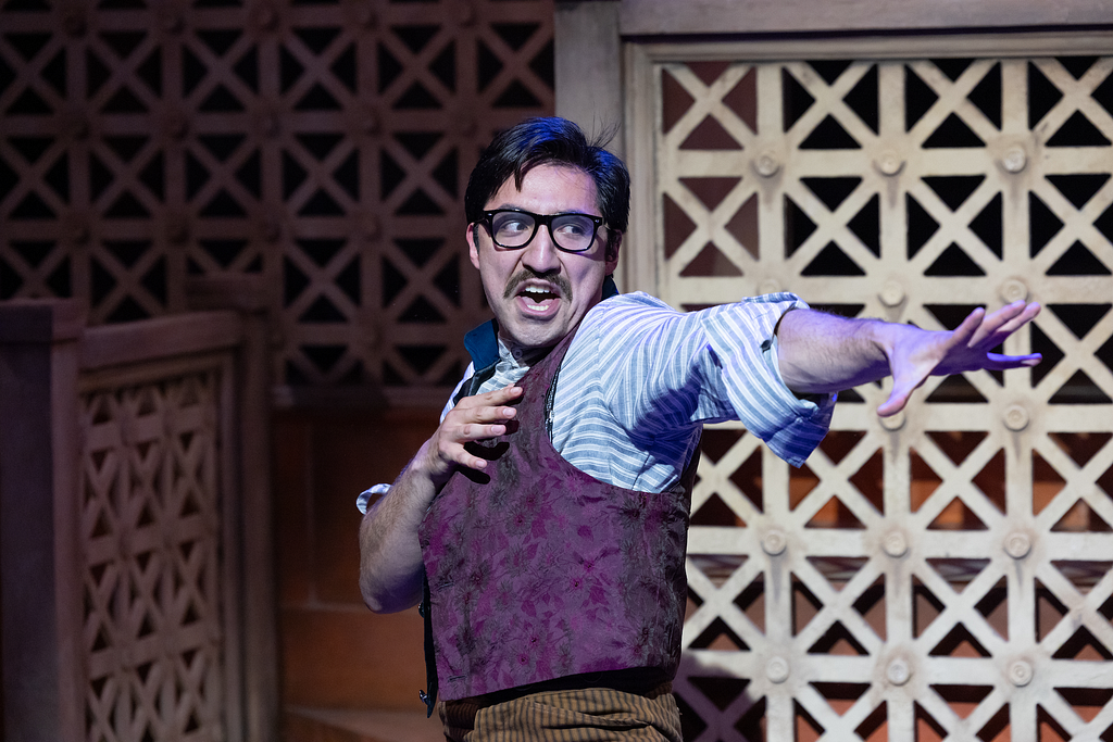 A man in a vest and black glasses clutches his heart and reaches out, performing dramatically.