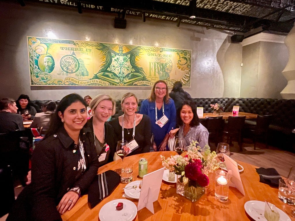 A photograph of five women with their TEDWomen conference badges on and smiling at their dinner table, with the author in the middle of the group.