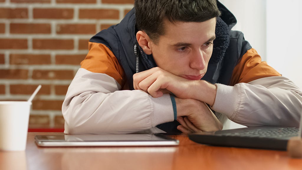Young man with his arms folded over a closed laptop staring into space with a sad look on his face.