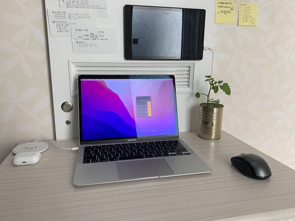 A work desk featuring a laptop, a mouse, and a tomato plant growing in an empty tomato can. Notes are pinned on the wall above the desk, and a tablet is mounted next to them.