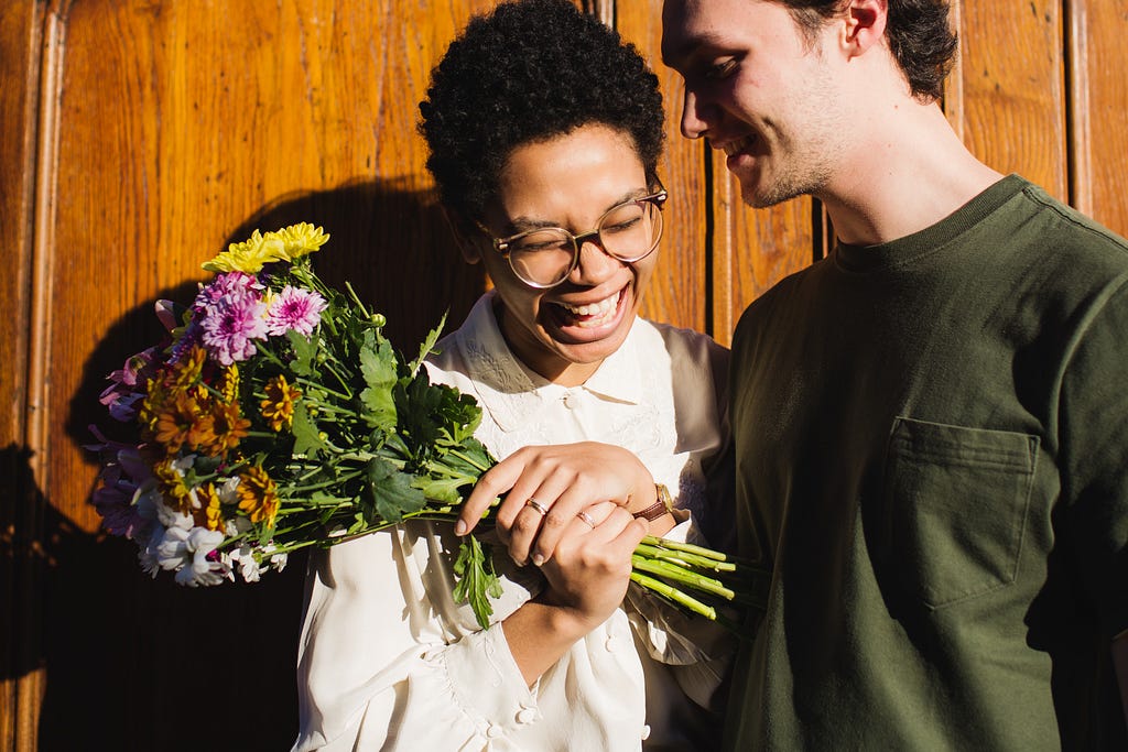 2 people standing next to each other. The woman is holding some flowers and smiling, while the man is leaning toward her and smiling