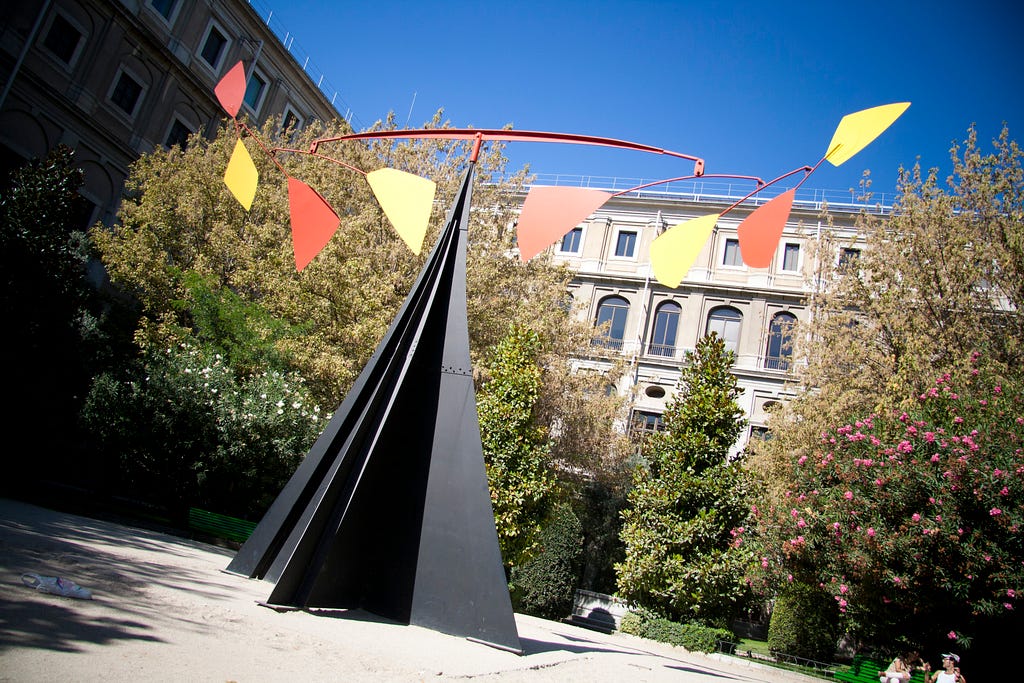 In a courtyard in front of the neoclassical facade of a building, where it is surrounded by healthy small green trees and flowering shrubs, a triangular black metal base supports a red bar to which eight smaller triangular panels are attached, alternating in color between red and yellow.