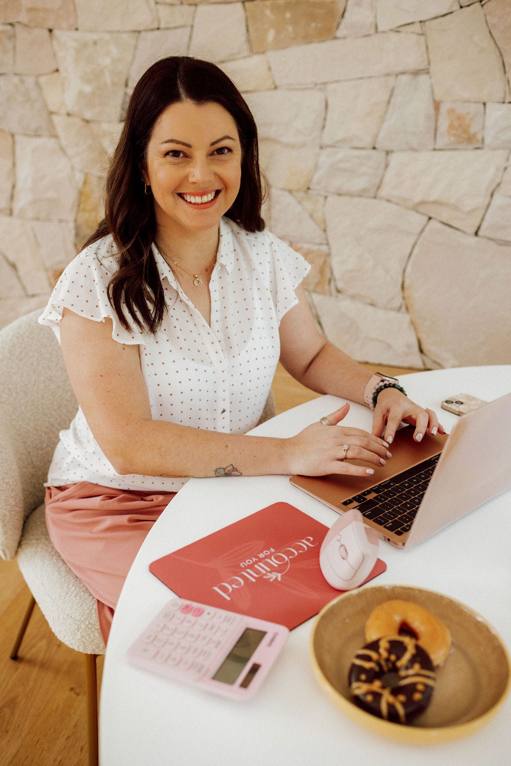 Lisa Turner in a white shirt, seated at a round table working on her laptop, with a calculator and plate of donuts in front of her.