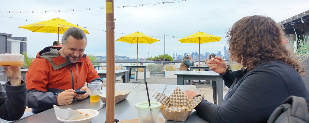 Two people seated at a patio table with drinks and takeout food at Marination in West Seattle, under yellow umbrellas with the Seattle skyline in the background on a cloudy day.