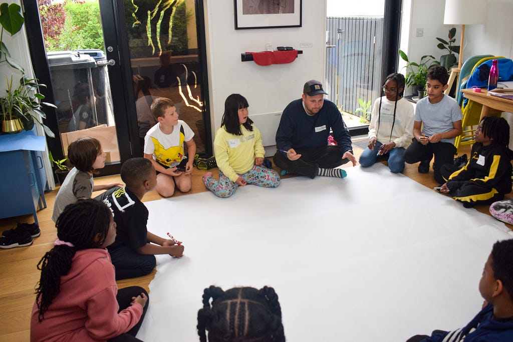 Polarbear sits with a group of children and young people — our YARD Youngers — gathered at YARD, in a circle around a large blank canvas. There are house plants, a large lamp and a pair of shoes in the background.