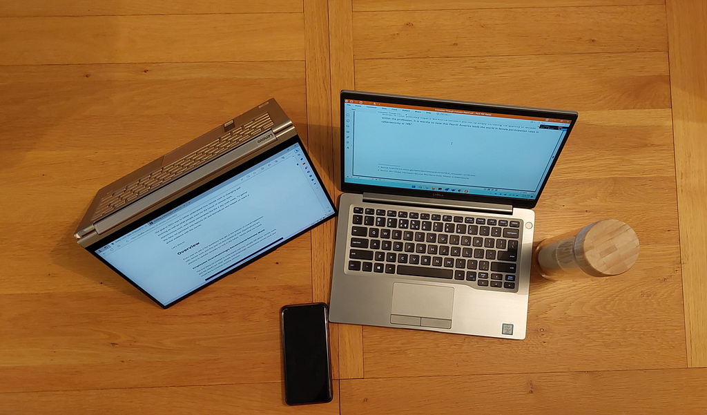 2 laptop computers (1 in tablet mode), a phone, and a coffee mug/bottle on a brown table