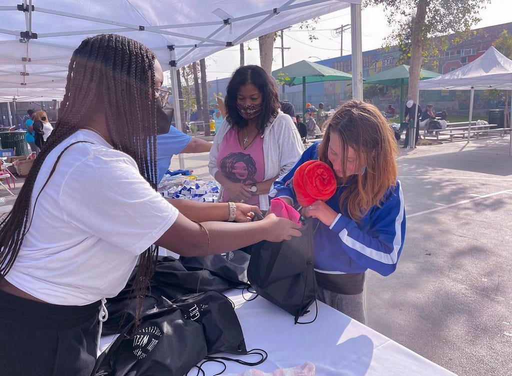 Teen girl with box braids wearing mask and white T-shirt handing kit to another teen girl wearing a blue jacket across a table on a sunny day.