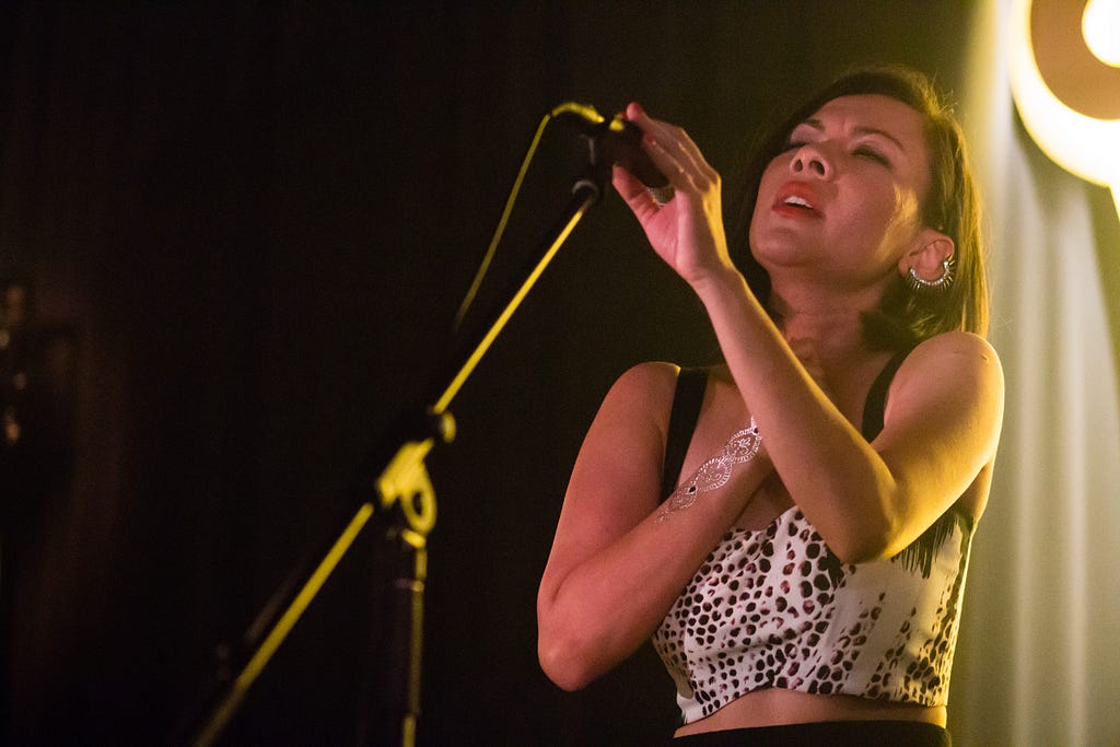 Rozella performing at Guinness Amplify. She is holding the microphone attached to the stand on her left hand, with her right hand on her chest.