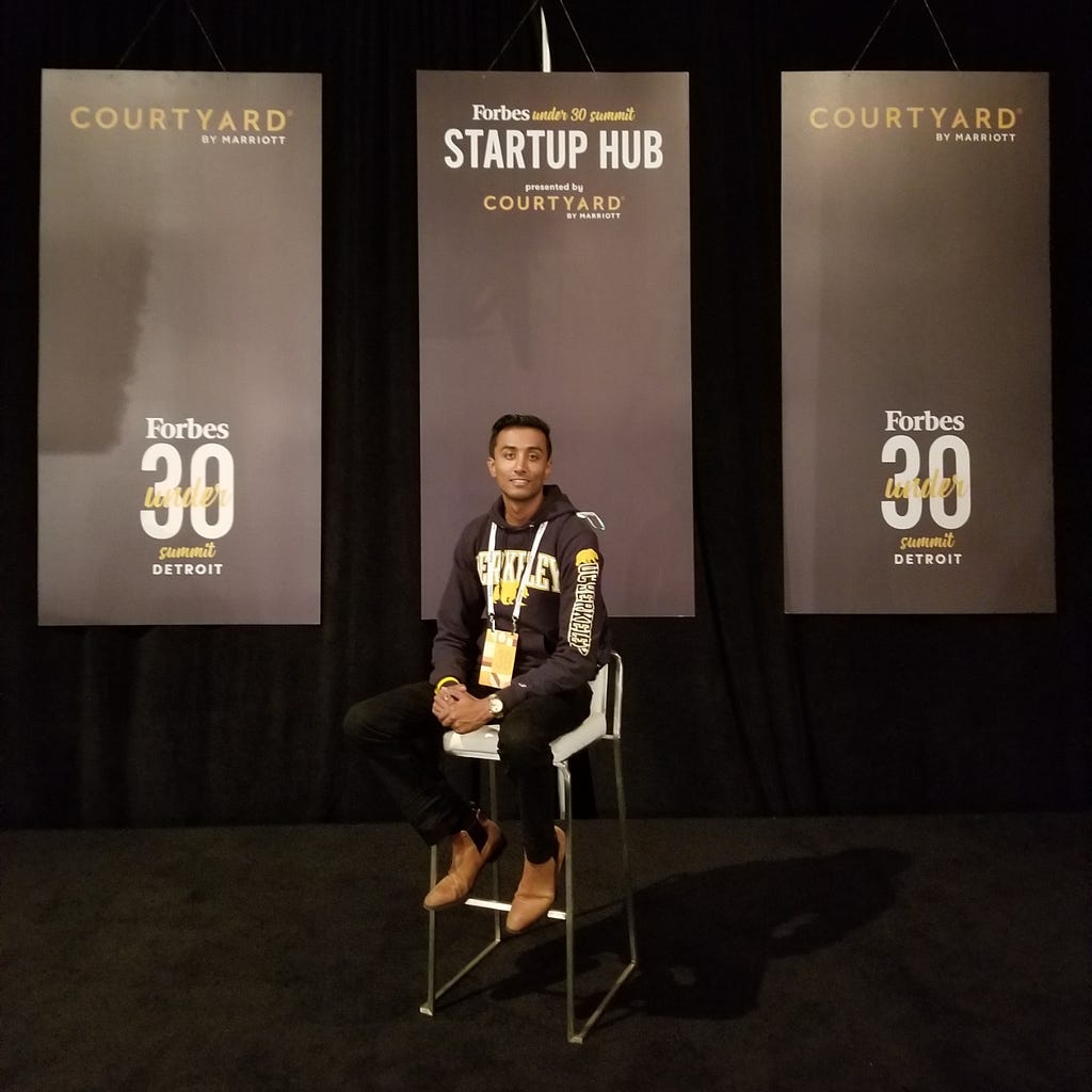 Ishmail sitting on a stool in front of three back banners with the Under 30 logo.