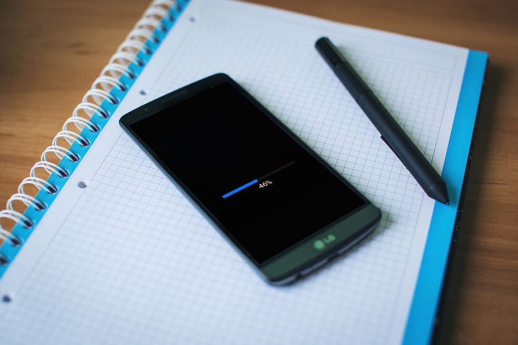 A black smartphone sits next to a black pen, on top of a white and blue notepad. The smartphone’s screen shows the loading bar at 46%.