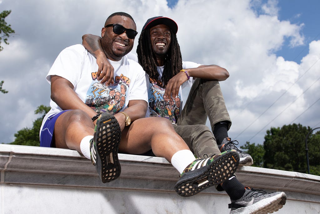Two black men sit on a ledge, smiling. The one on right, H.D. Hunter, has his arm around the other, Olujide Lawal. They are wearing matching white t-shirts and sneakers. Olujide is wearing black sunglasses. H.D. Hunter has on a baseball cap.