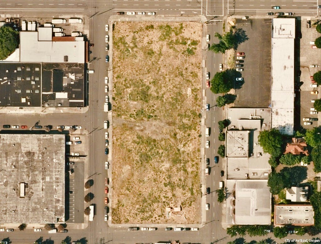 An aerial view of a two-block long empty field running north to south, buildings around it across every street.