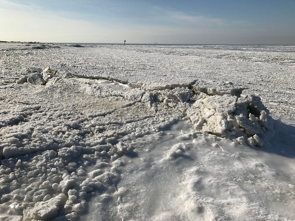 This photo shows Delaware Bay completely covered in a sheet of thick, white ice. Toward the center of the image, the ice is cracking and raising. Underneath that feature is an oyster-reef breakwater, though it’s not visible in the image.