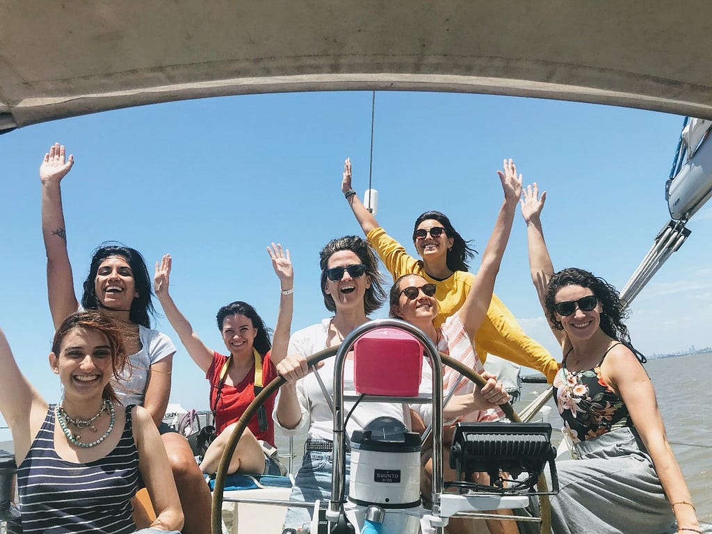 A group of woman are smiling on a boat on a clear day, their hands in the air.