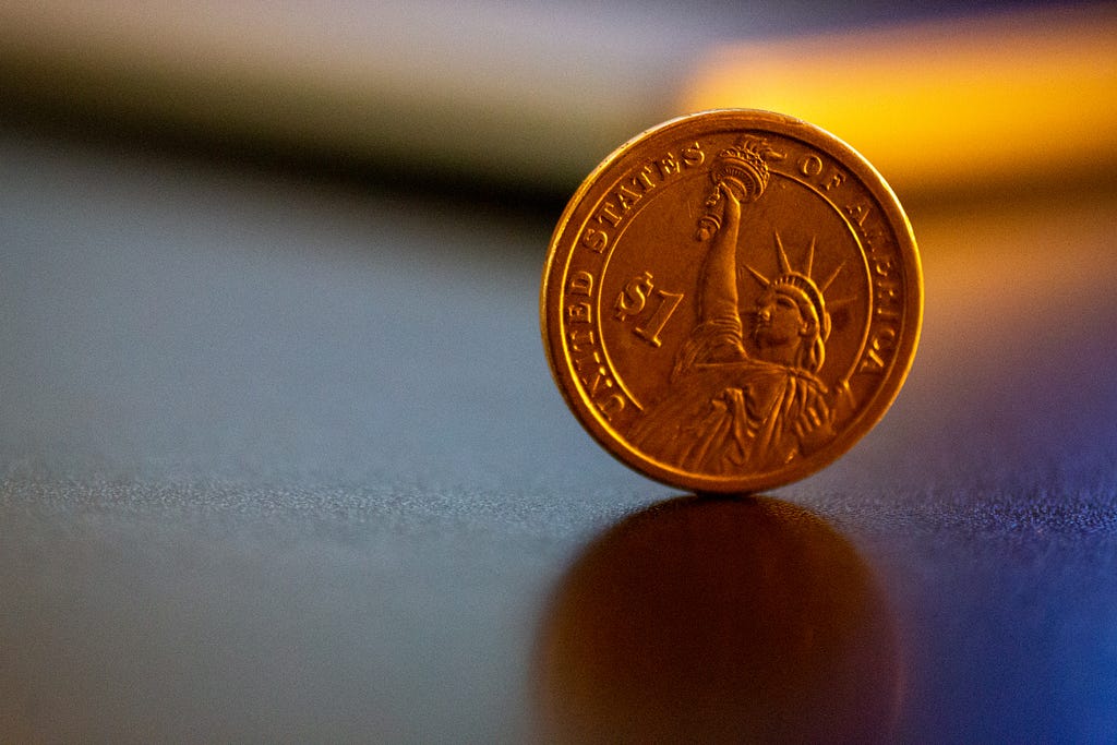 A close-up photo of a United States dollar coin. The coin is standing upright on a desk in full focus, whilst the surroundings are blurred.