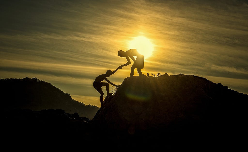 A person helping another person to get to the top of the hill at the end of the day with the sun dimming