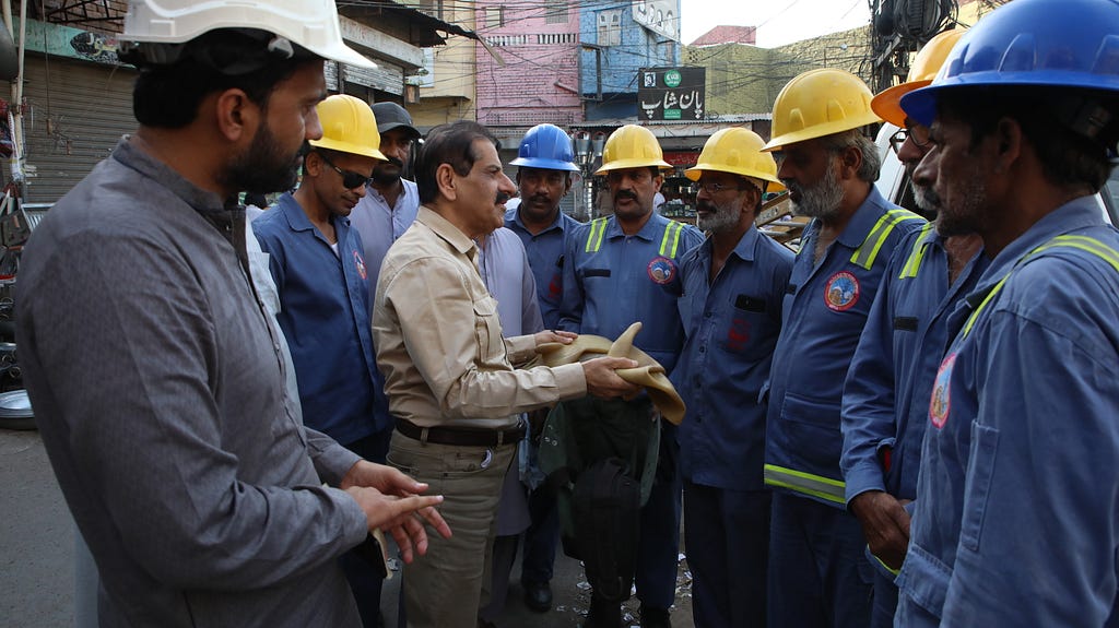 A group of men dressed in work uniforms and hardhats listen to another man speaking to them.
