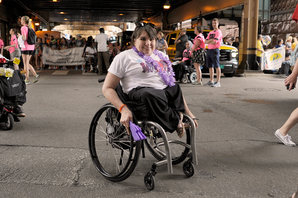 Karen wears a casual outfit and a purple lei, holding a purple noisemaker. She has short brown hair and uses a wheelchair.