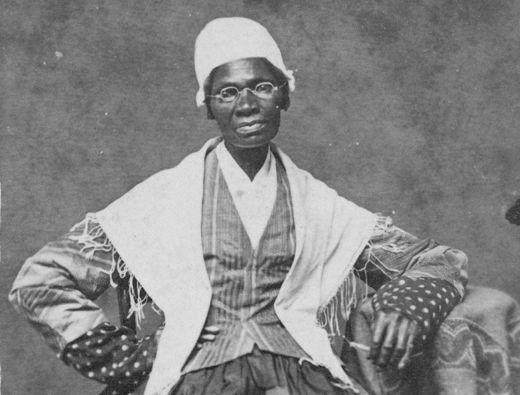 Truth sits posed while seated with a white shawl over her dress and a headwrap, with glasses on.