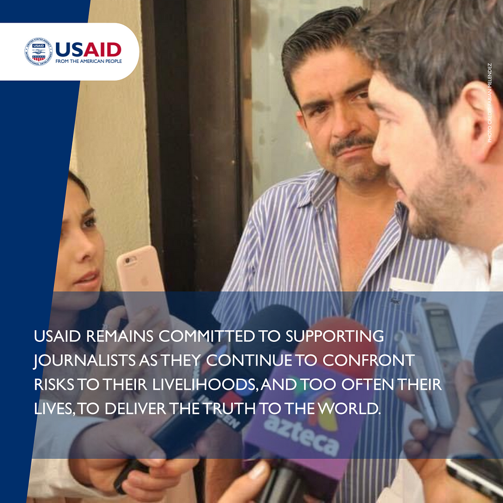 A man speaking into two microphones is interviewed by two journalists, with one of the journalists holding up a smartphone to record the interview. A statement below the image reads: USAID remains committed to supporting journalists as they continue to confront risks to their livelihoods, and too often their lives, to deliver the truth to the world.