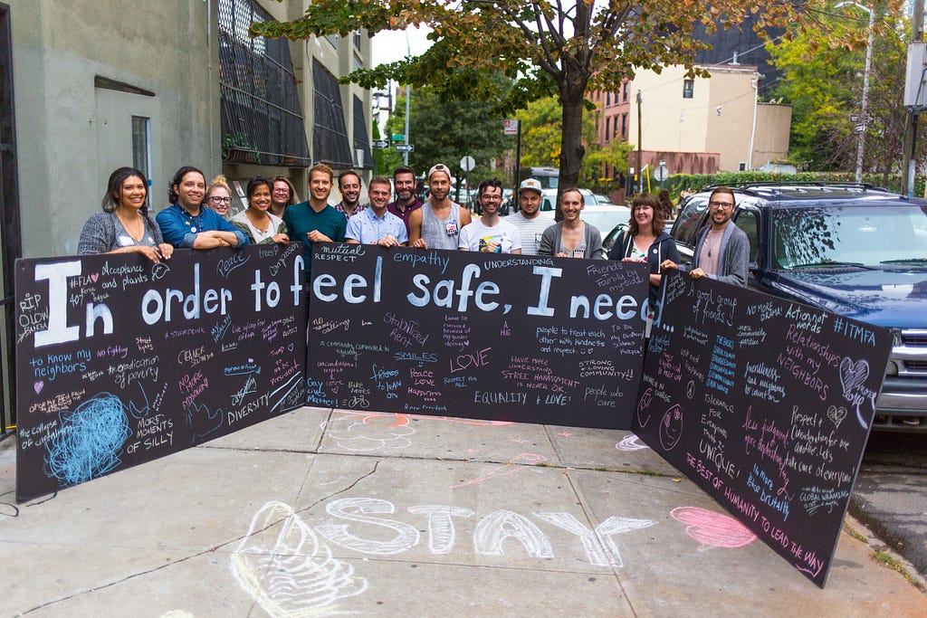 A sizable group of people pose behind a chalkboard that says “In order to feel safe, I need…” including answers that people have written in.