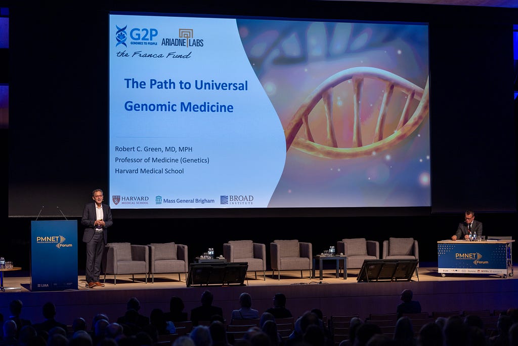 Dr. Robert C. Green stands at a podium in front of a screen displaying a title slide: “The Path to Universal Genomic Medicine.”