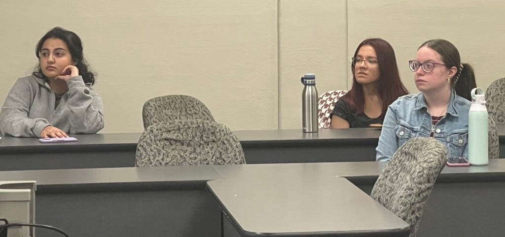 Three students sitting in rolling chairs in a classroom listening to a presentation.