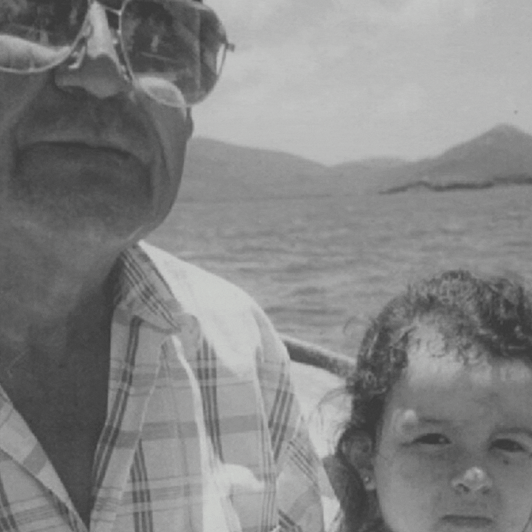 Gif with a black and white portrait of Thania Betancourt Alcazar as a child sitting next to her late grandpa Manuel on a boat. Fading into the front is a colored image of Thania’s hand holding her grandpa’s bumpersticker that says I heart democracy in spanish