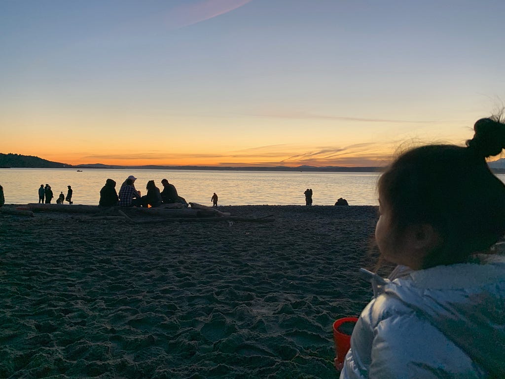 A photo of a young child in profile wearing a silver hooded jacket with a bun. The child is looking out at some friends on the beach enjoying the winter sunset over the Olympic Mountains.