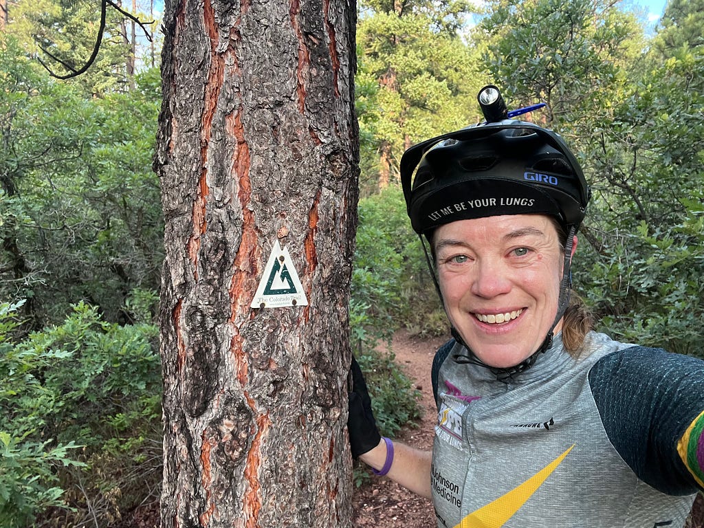 The author, Patty George, smiling in her Team PHenomenal Hope uniform and bicycle helmet, standing next to a tree with the Colorado trail logo. She is wearing a cycling cap with the words “Let Me Be Your Lungs”