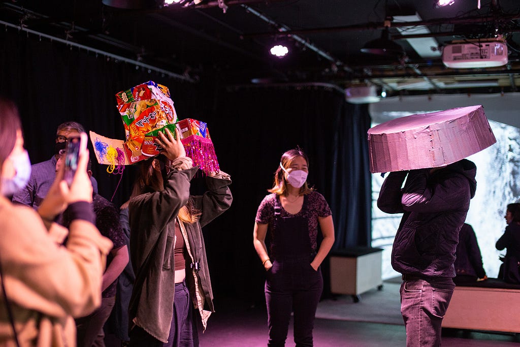 A group faces each other in the dark CultureHub studio, one wearing a cardboard washbasin on their head, while another wears a multicolored Korean crown covered with snack wrappers. Ann looks on from the back, wearing a mask.