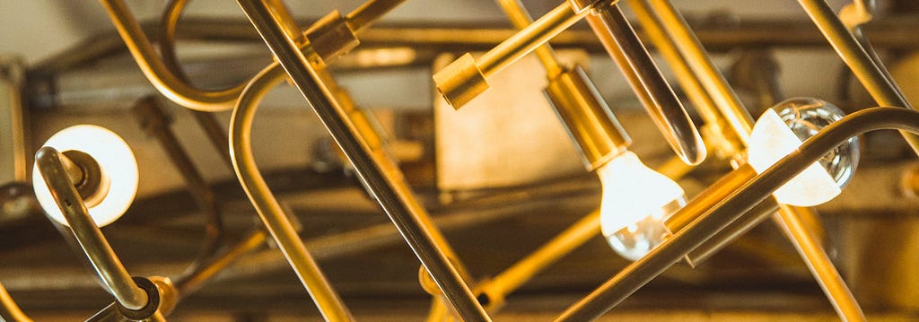 Golden metal pipes connect softly glowing lightbulbs in an complex, angular network.