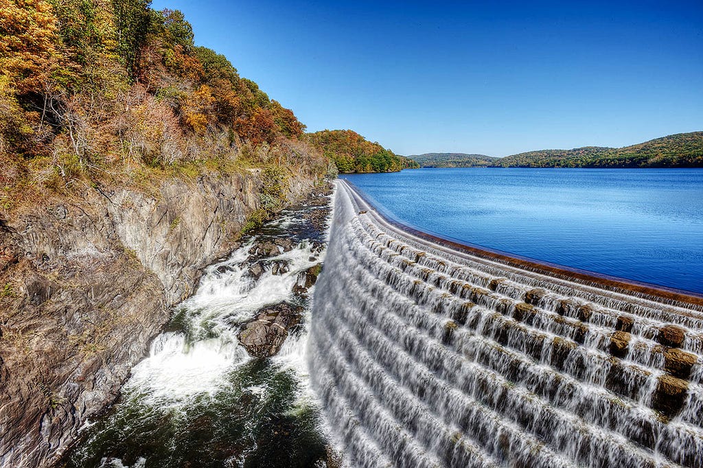 A scenic view of a stone dam with cascading water flowing over its spillway into a calm lake below. Surrounding the dam are lush green trees.