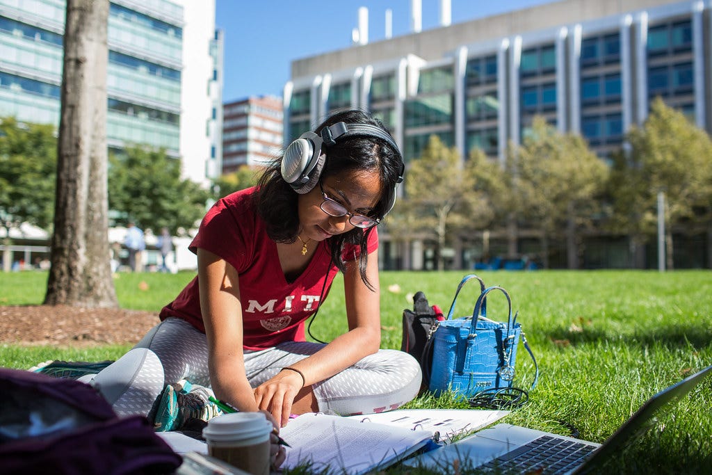 Female student with headphones sitting on grass, writing in a notebook in front of a laptop.