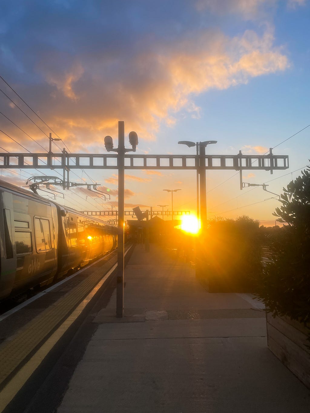 A train station, with a train on the left-hand side, overhead lines and the sun setting