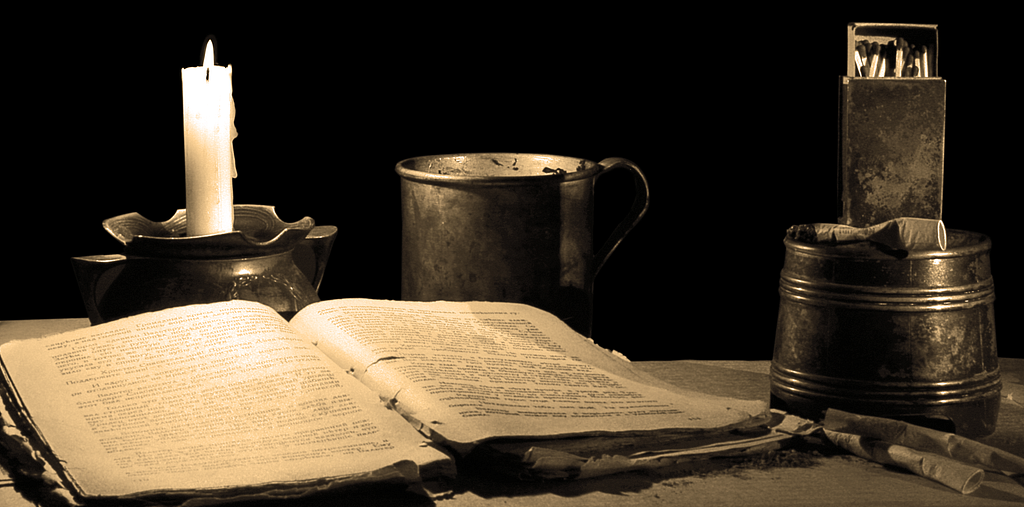 Tampo de uma mesa empoeirada com uma vela à esquerda, um livro antigo aberto e uma caneca de metal