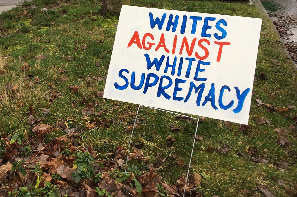 Hand-painted sign reading WHITES AGAINST WHITE SUPREMACY in red, white, and blue, displayed in a front yard.