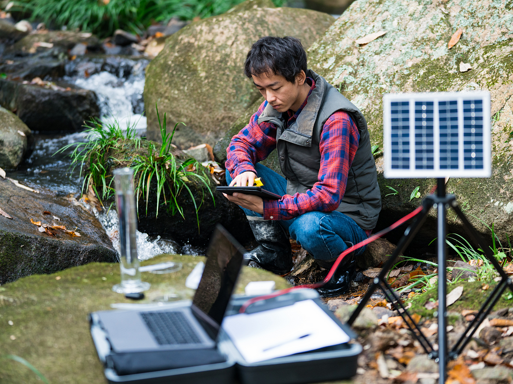 A field scientist kneels by a stream, using a tablet for data collection. He is surrounded by scientific equipment, including a solar panel setup and a laptop.