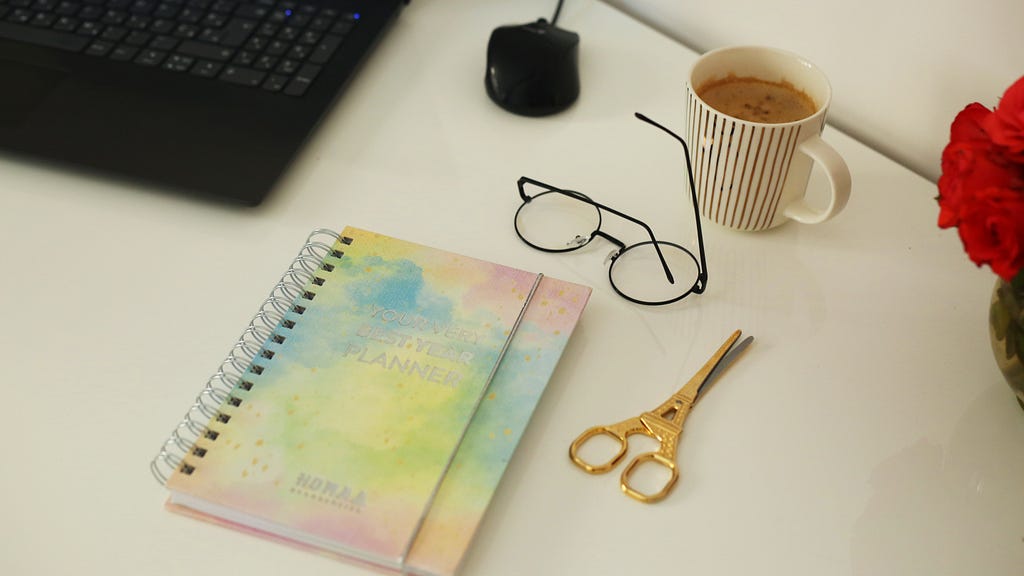 Photo of a computer keyboard and mouse, spiral bound journal, scissors, glasses, cup of coffee.