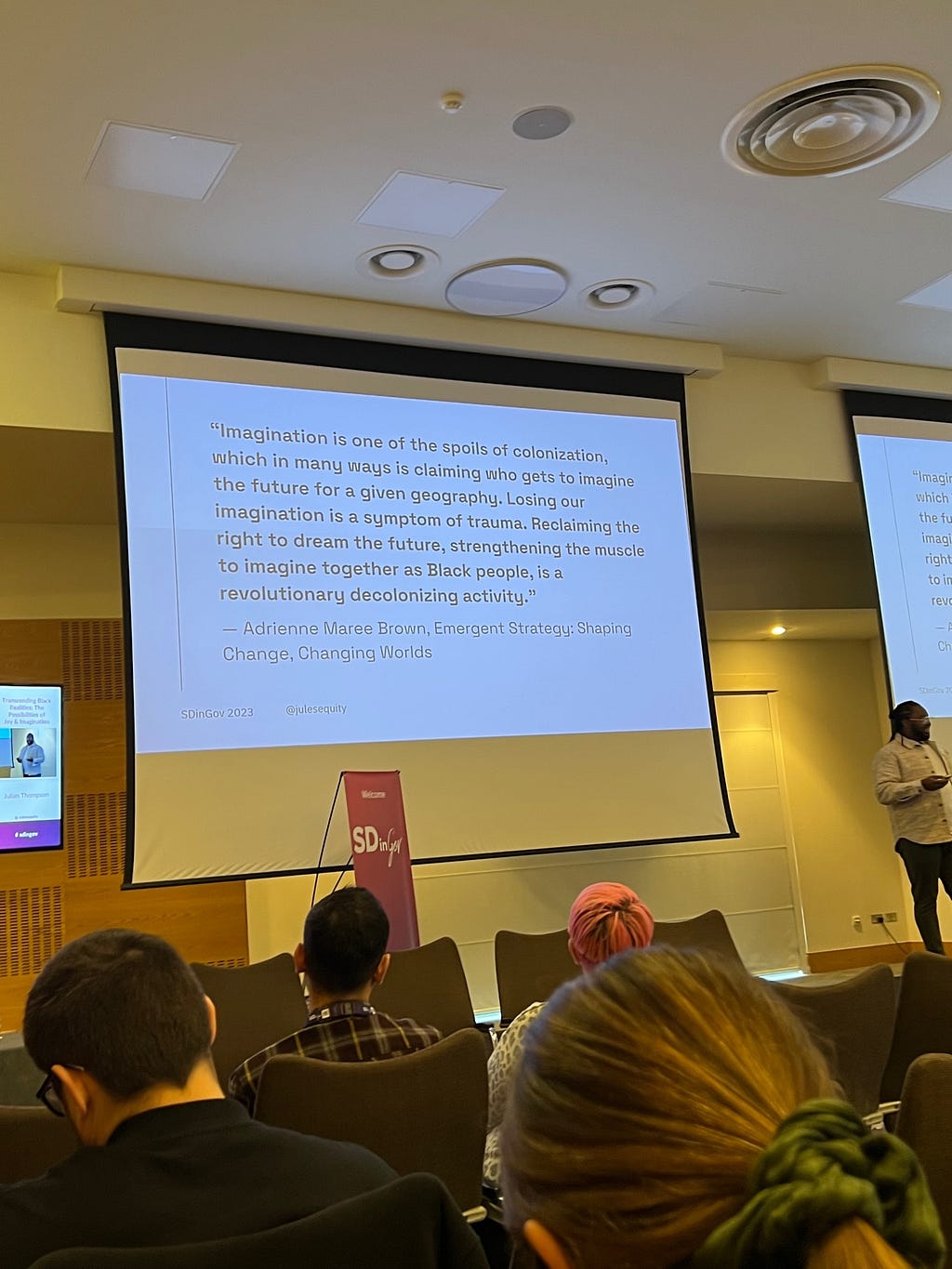 Julian Thompson stands on a stage at a conference, in front of a slide that reads: ““Imagination is one of the spoils of colonization, which in many ways is claiming who gets to imagine the future for a given geography. Losing our imagination is a symptom of trauma. Reclaiming the right to dream the future, strengthening the muscle to imagine together as Black people, is a revolutionary decolonizing activity.” The quote is by Adrienne Maree Brown