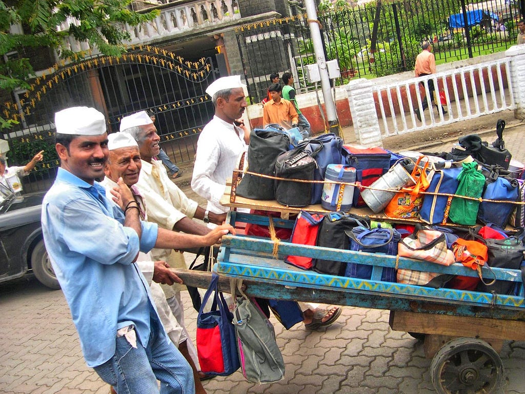 Four Indian men, all wearing white cloth caps push trailers loaded with lunch tins and bags.