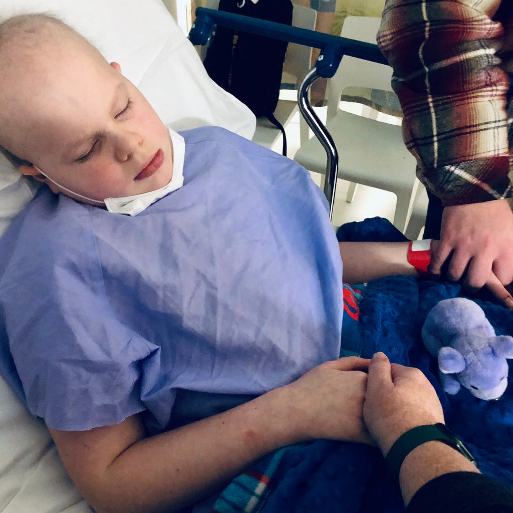 A boy fighting cancer, laying in a hospital bed with his parents holding his hand.